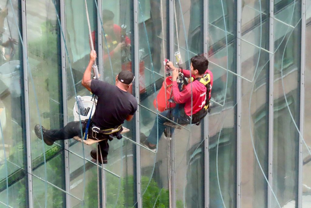 Two professional window cleaners working on a tall glass building using suspended harnesses.