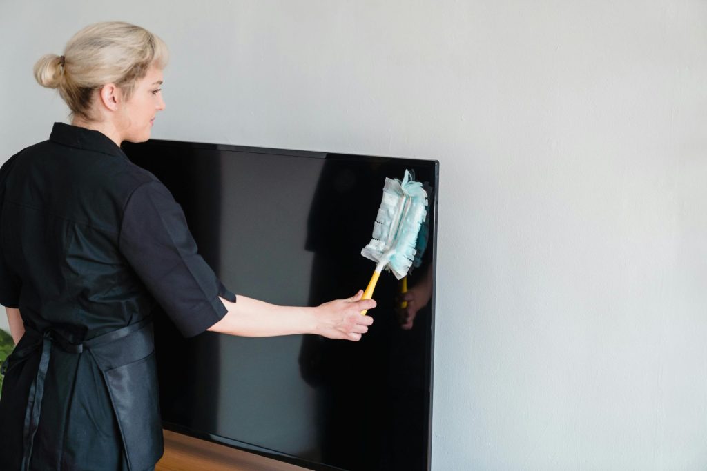 A female housekeeper performing dusting on a television screen indoors.